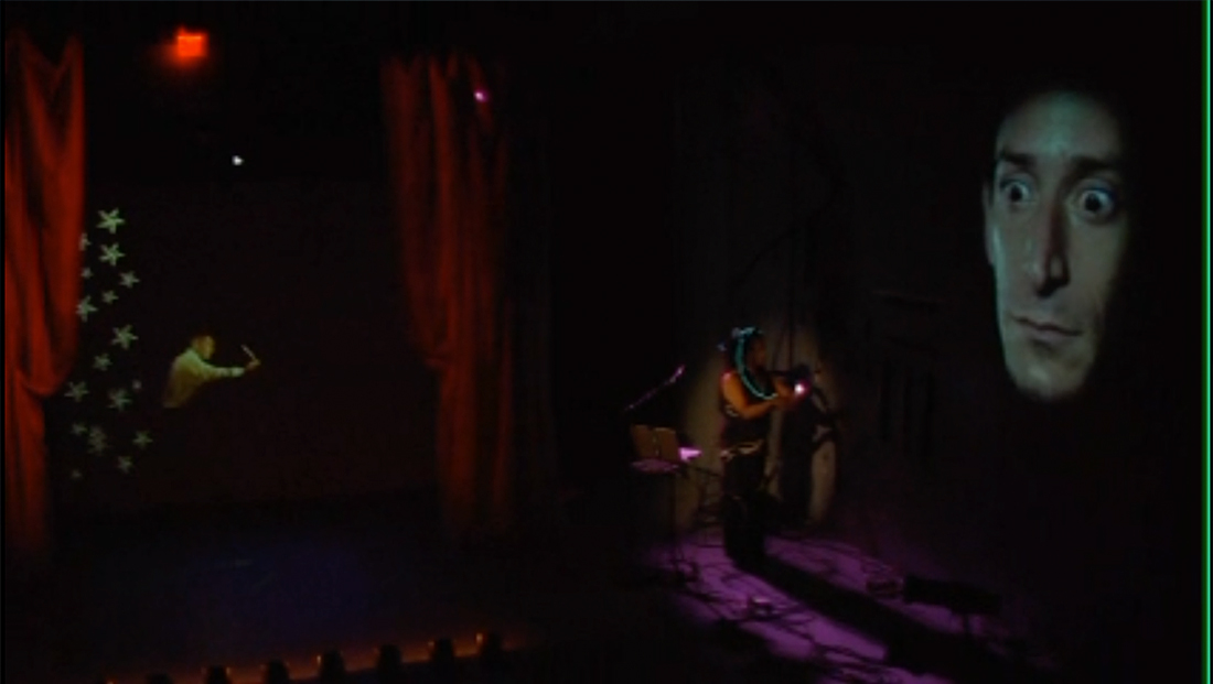 Still from the video POE (…AND THE MUSEUM OF LOST ARTS) , projected face looming over a dark stage with two actors and red curtains