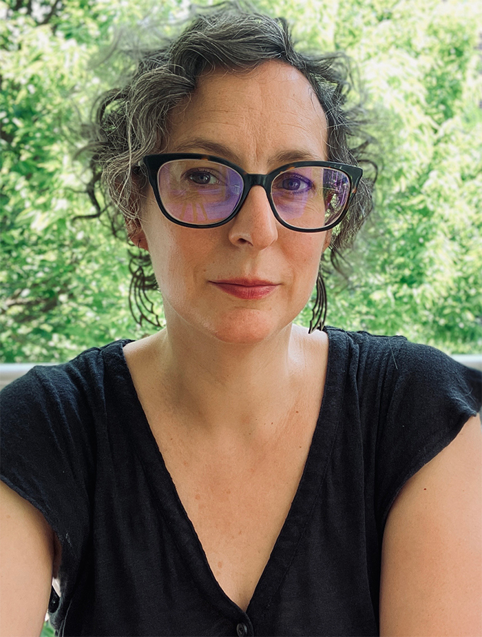 headshot of Stefanie Koseff, seated outdoors in front of green foliage wearing black framed glasses and a black t-shirt